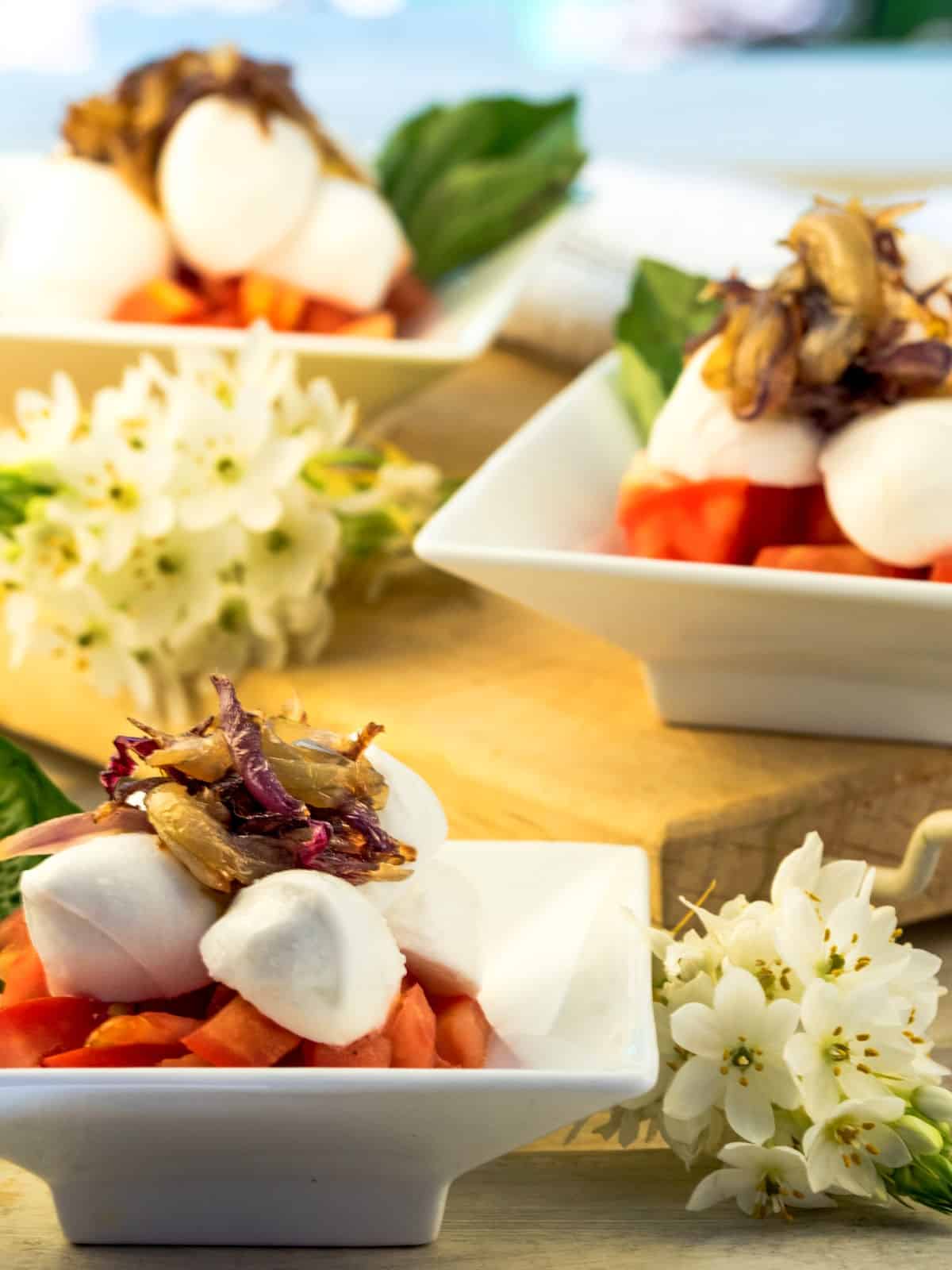 Bocconcini Caprese with caramelized onions on serving dishes and flowers in the background.