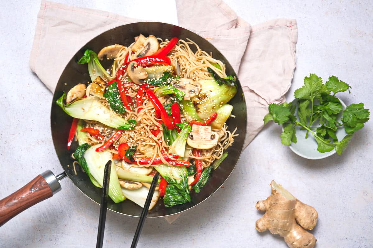 Bok choy stir fry with noodles in frying pan on light background. Chopsticks, ginger root and cilantro on the side.