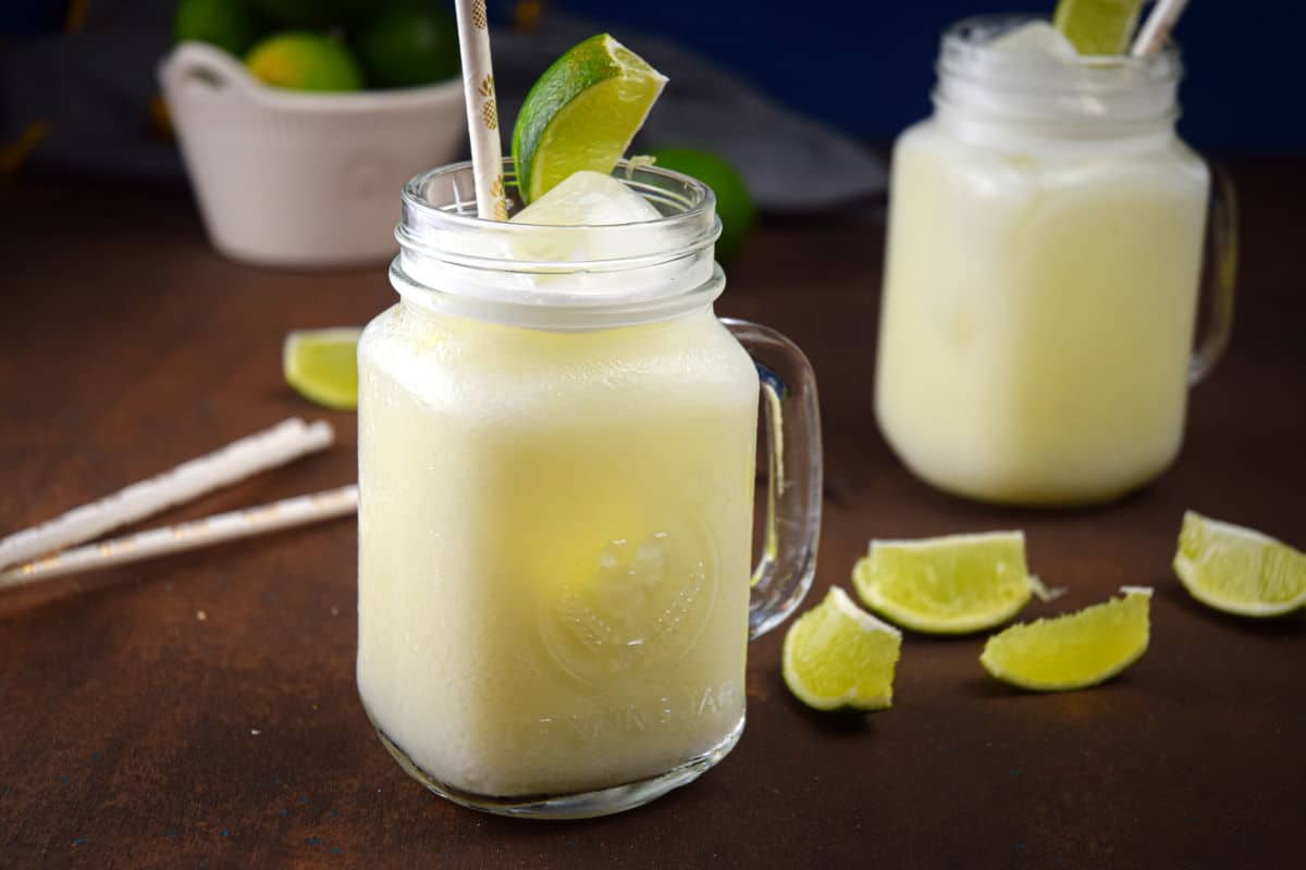 Brazilian lemonade in glass jars with lime wedges and paper straws, limes in a bowl in the background.