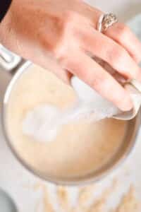 A woman's hand holding a small bowl of sugar over a pot.