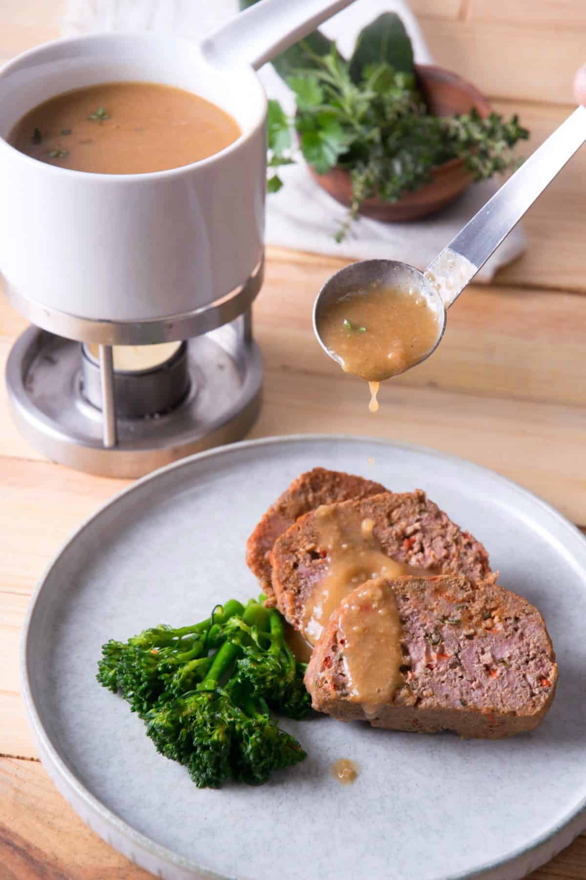 Gravy dripping over plate of meatloaf and broccoli, gravy warmer in the background.