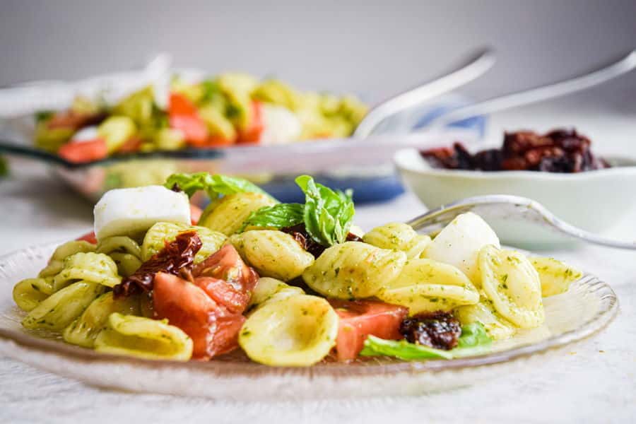 Bruschetta pasta salad on clear glass plate, white background and small bowl of sun-dried tomatoes in the background.