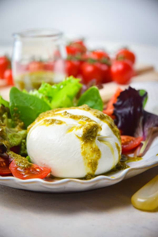 Burrata salad on a white plate, tomatoes and pesto in the background.
