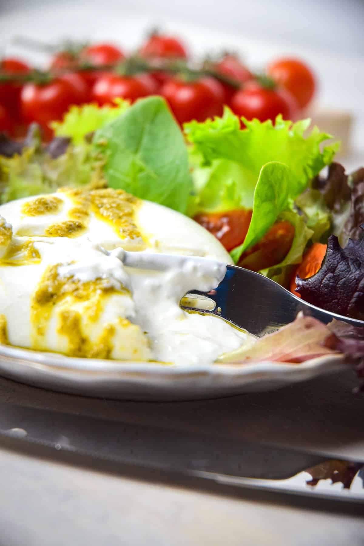 Burrata salad on a white plate with a fork, tomatoes in the background.