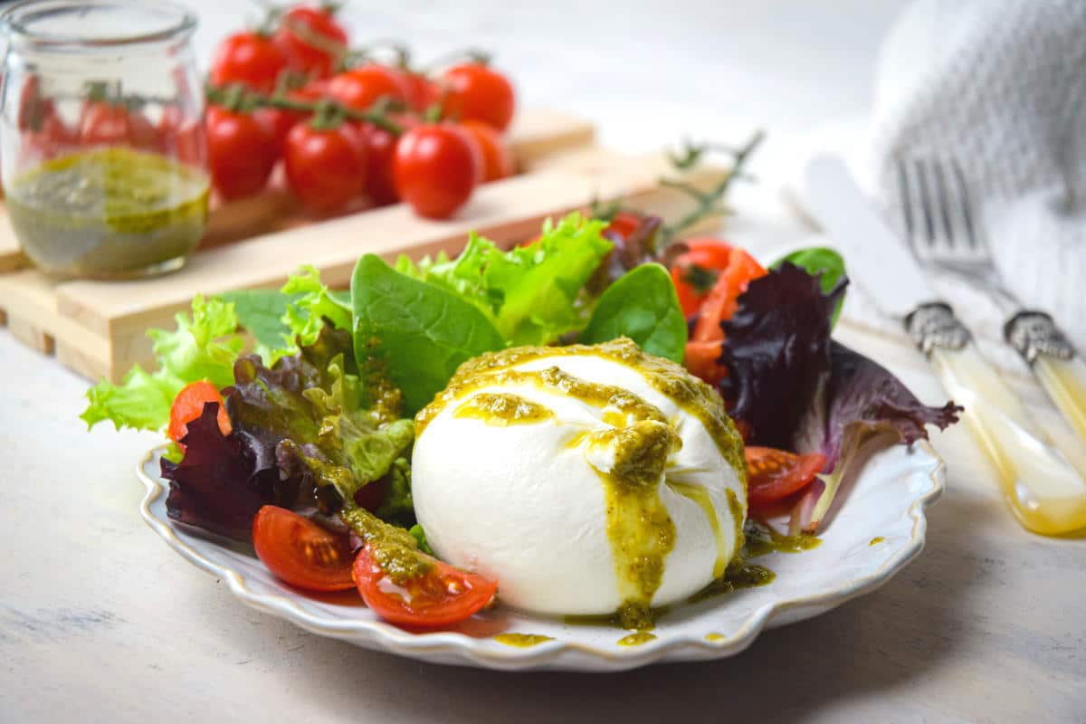Burrata salad on a white plate, tomatoes and pesto in the background.