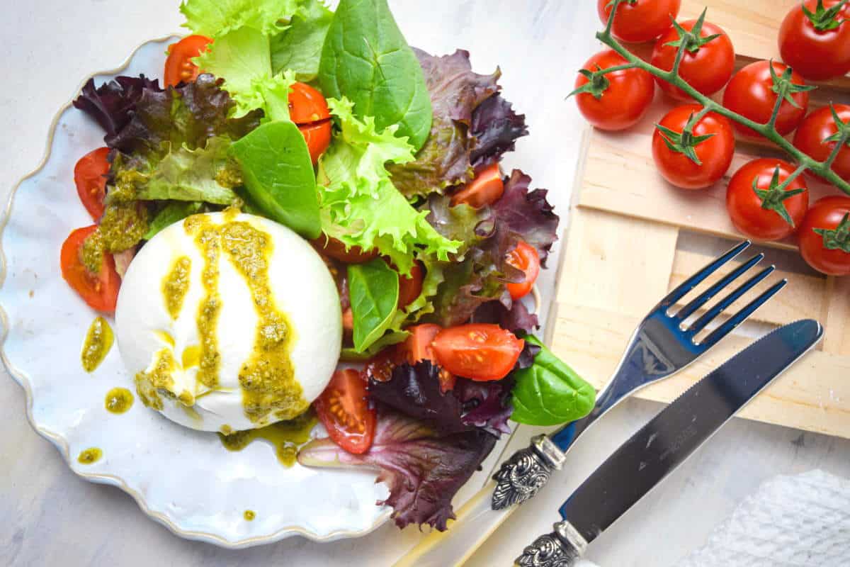 Burrata salad on a white plate, tomatoes, pesto and cutlery in the background.