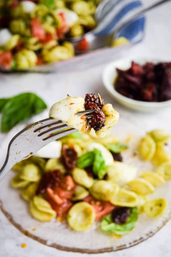 Caprese pasta salad with a fork on clear glass plate, white background, sun-dried tomatoes in a small bowl on the side.