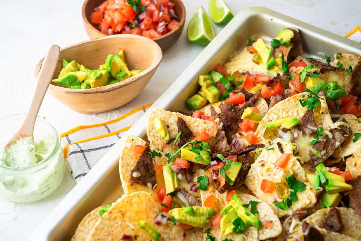 Carne Asada Nachos in a pan, with lime wedges, tomatoes, avocado and cilantro dip on the side.