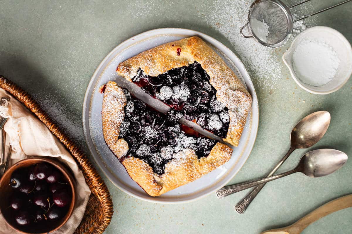 Cherry galette with icing sugar on white plate. A small bowl of cherries and icing sugar on the side.