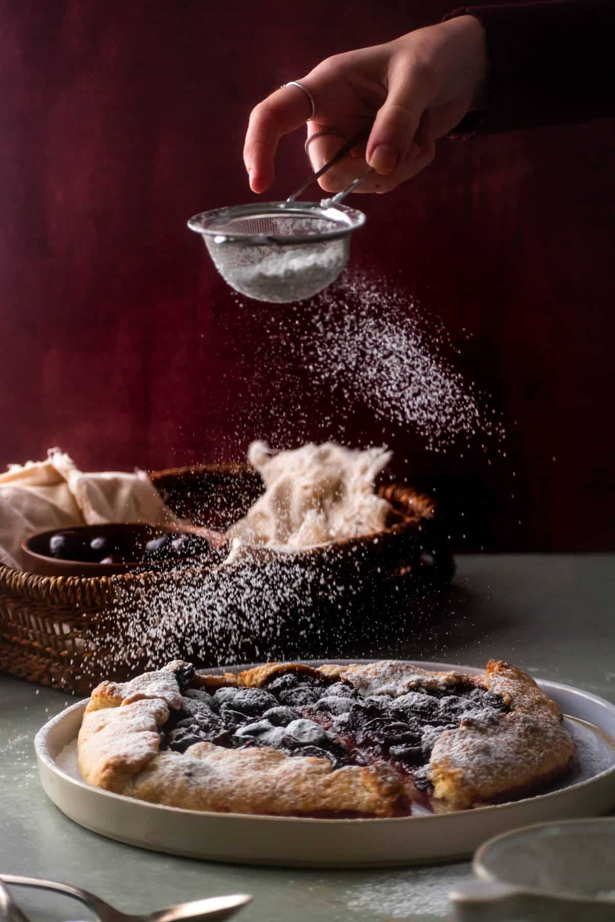 Cherry galette with icing sugar sprinkled on top, on white plate. Dark red background.