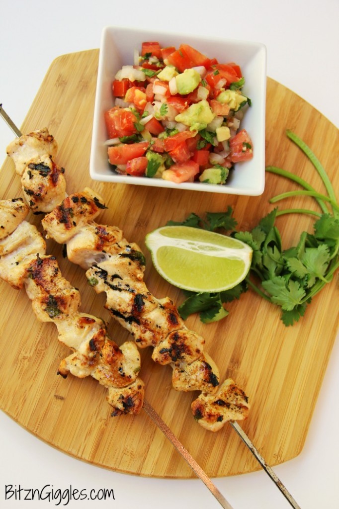 Chicken skewers, cilantro, lime wedge and bowl of salsa on wooden board.