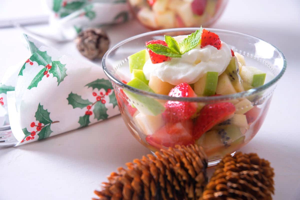Christmas fruit salad with whipped cream in a small glass bowl, pine cones and Christmas napkin on the side.