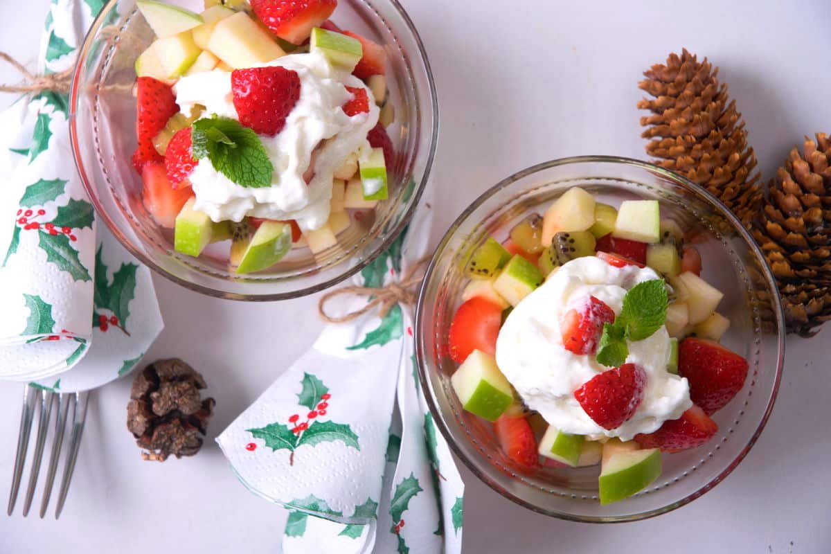 2 glass bowls with Christmas fruit salad. Pine cones, fork and Christmas napkins on the side.