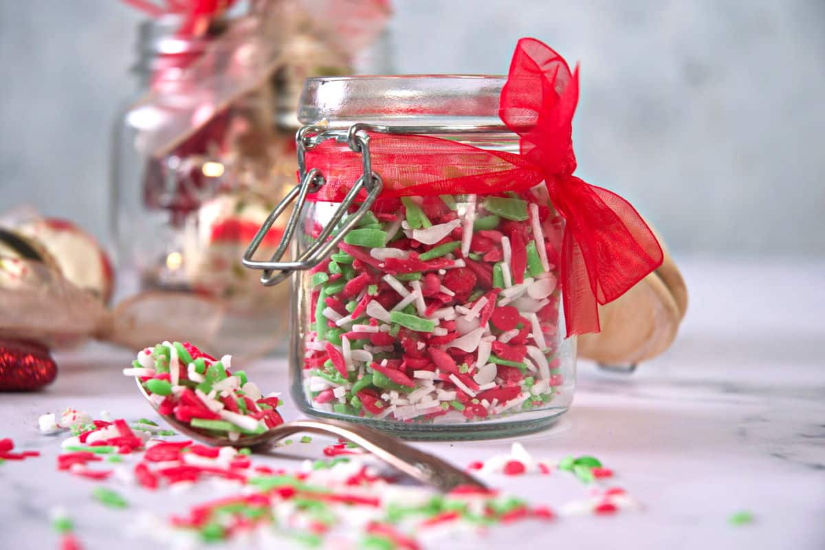 Red, green and white sprinkles in a small jar with a red ribbon and a spoon on the side.