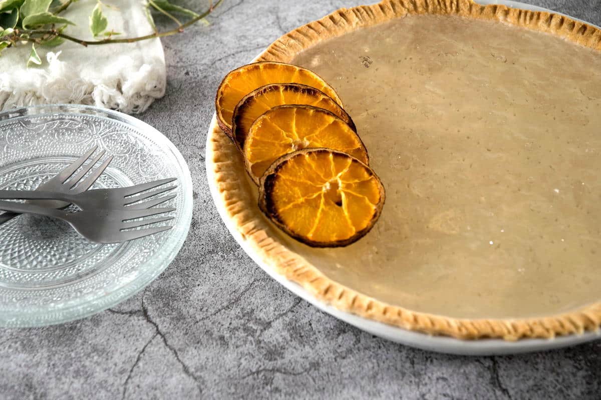 Clear pumpkin pie with dehydrated orange slices, forks and glass plates stacked on the side.