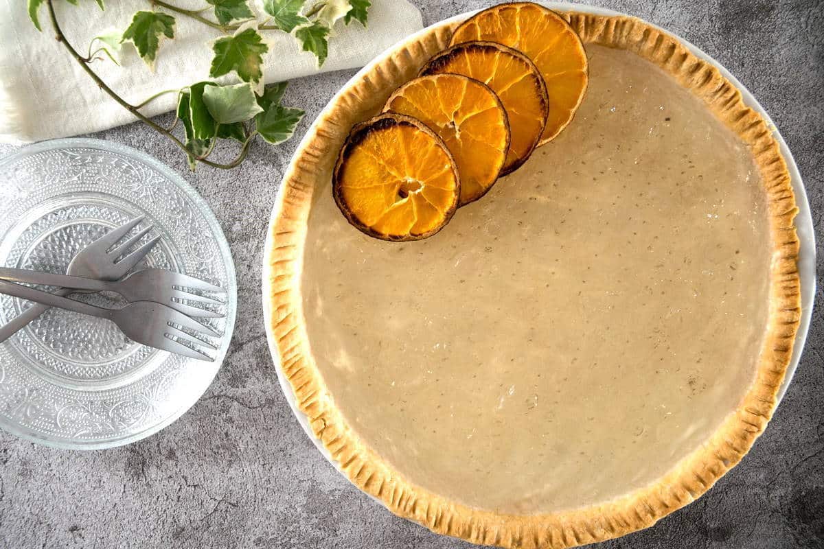 A pie with a golden crust and three dried orange slices on top is placed on a gray surface. Beside the pie, there is a clear glass plate with elegant patterns, two forks, a linen napkin, and some green ivy leaves.