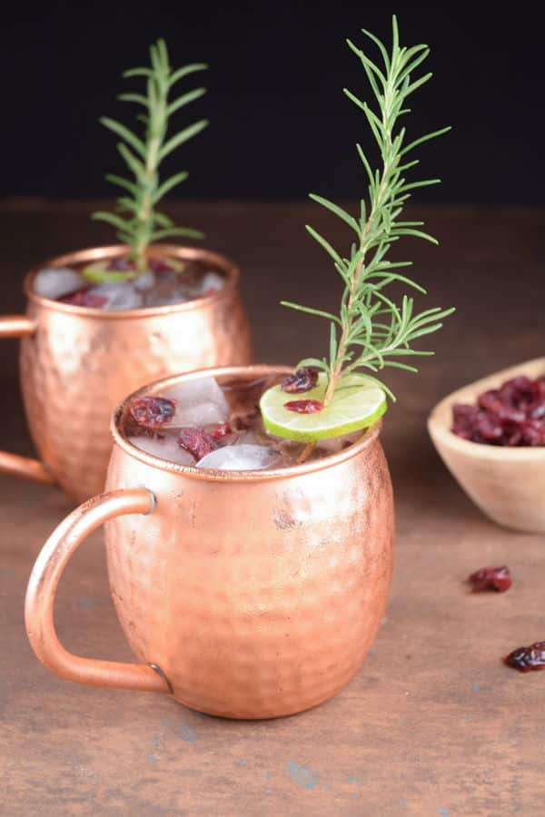 Cranberry Moscow Mule in copper mugs, garnished with cranberries, rosemary and lime slices.