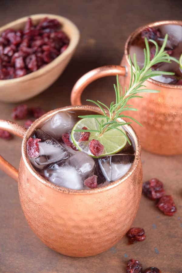 Cranberry Moscow Mule in copper mugs, garnished with cranberries, rosemary and lime slices.