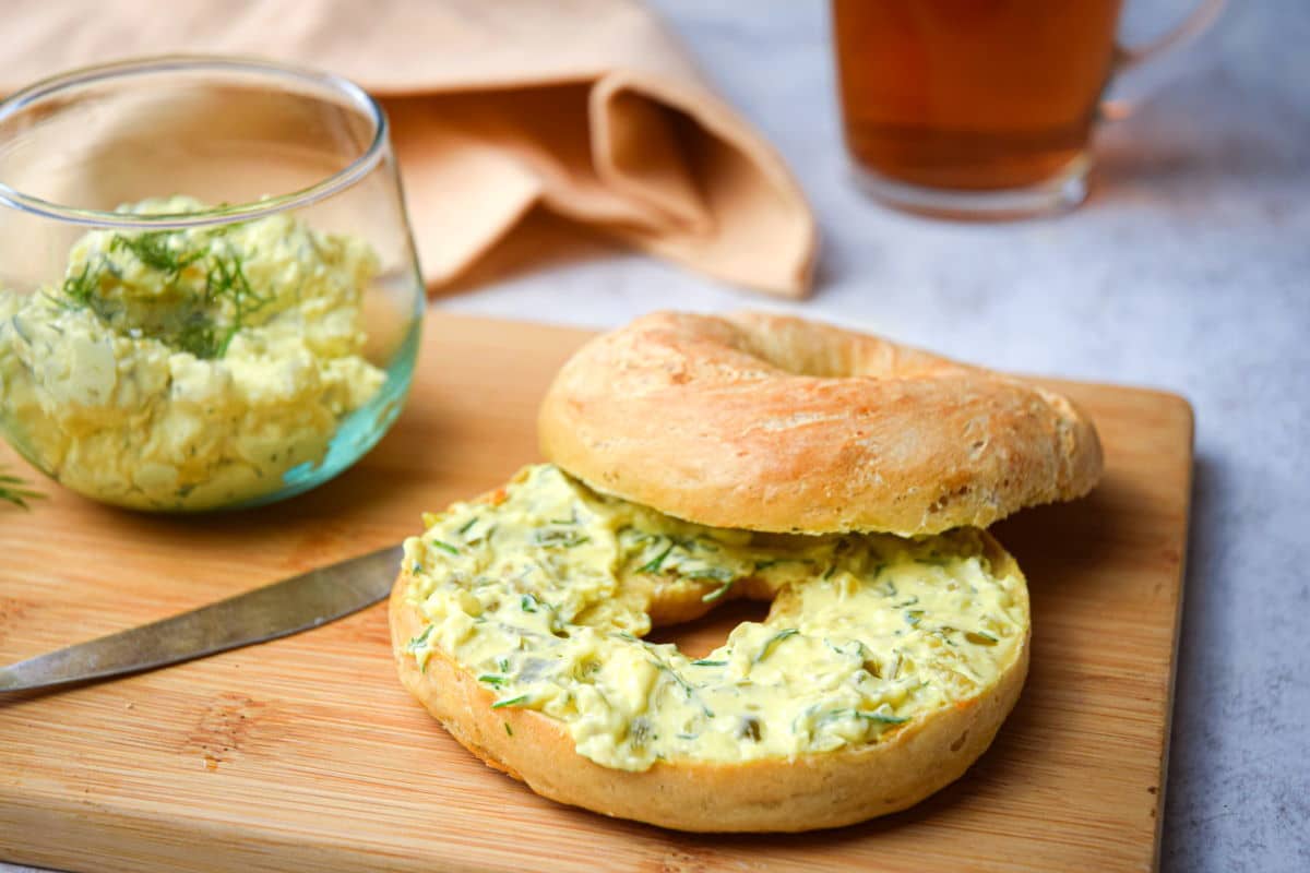 Dill pickle cream cheese spread on a bagel on wooden cutting board.