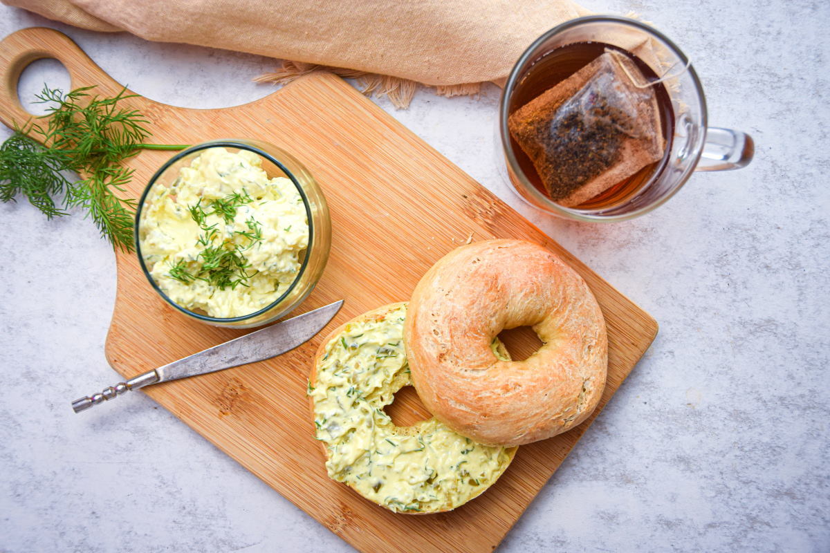 Dill pickle cream cheese spread on a bagel on wooden cutting board, a mug of tea on the side and fresh dill.