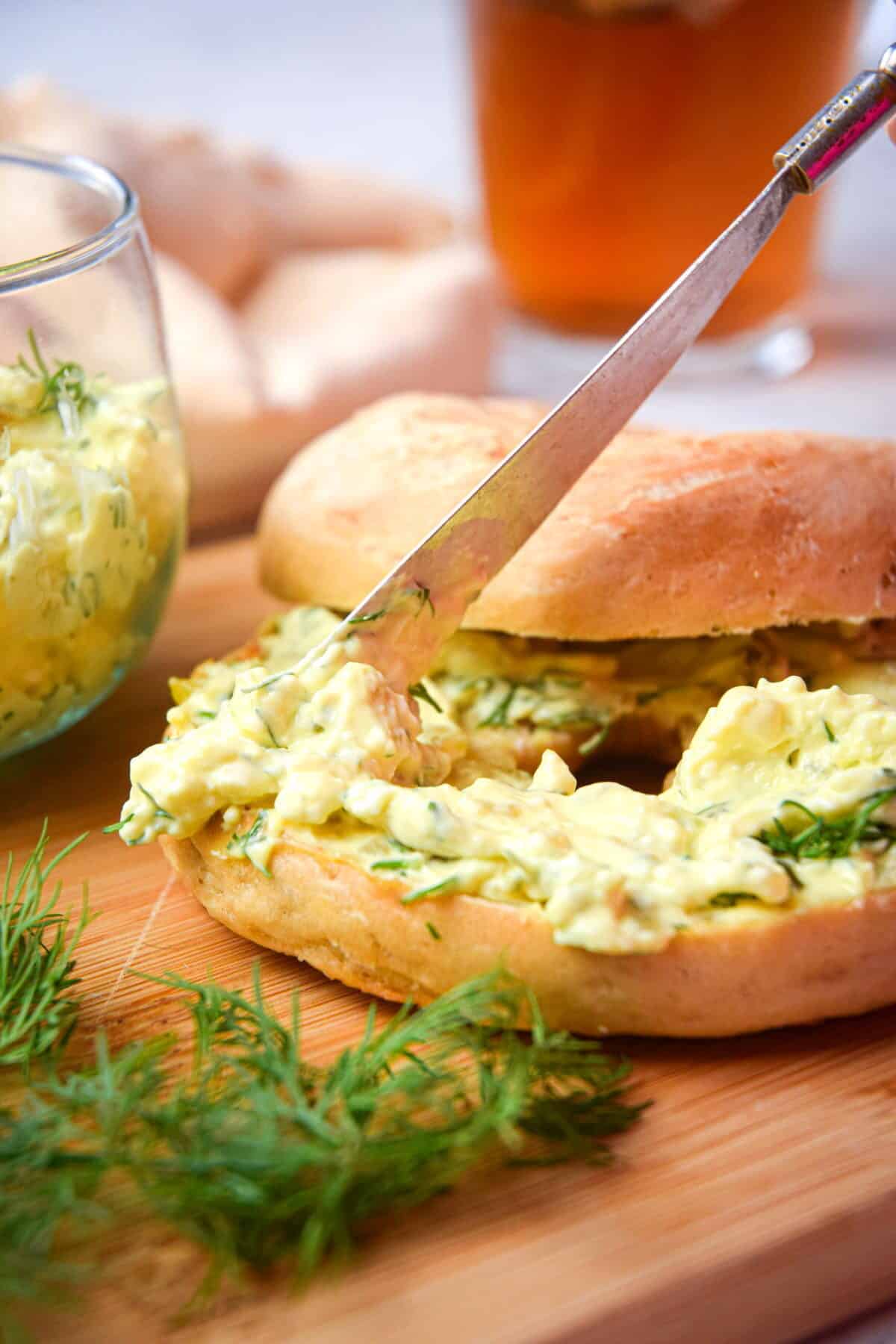 Dill pickle cream cheese spread on a bagel with a butter knife on wooden cutting board.