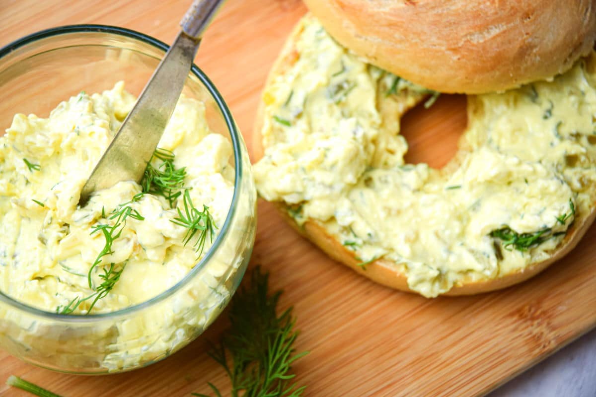 Dill pickle cream cheese spread in a small glass dish with a butter knife, a bagel with spread beside it on cutting board.