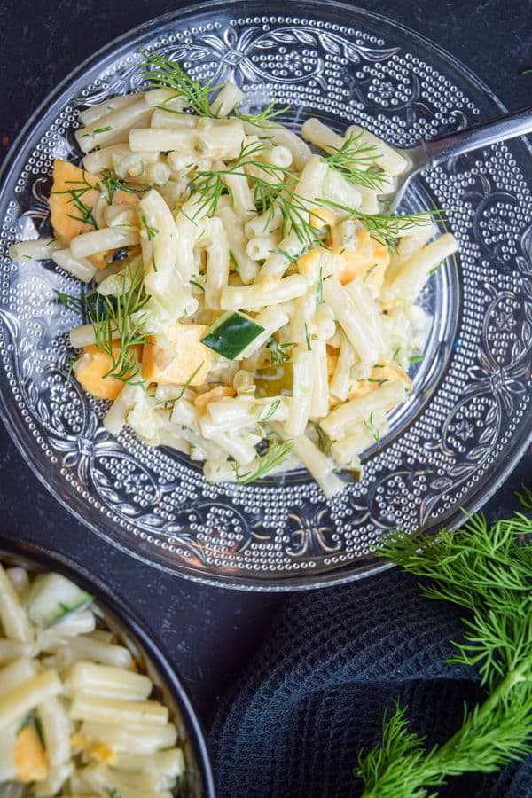 Dill pickle pasta salad garnished with fresh dill on a clear glass plate, dark background.