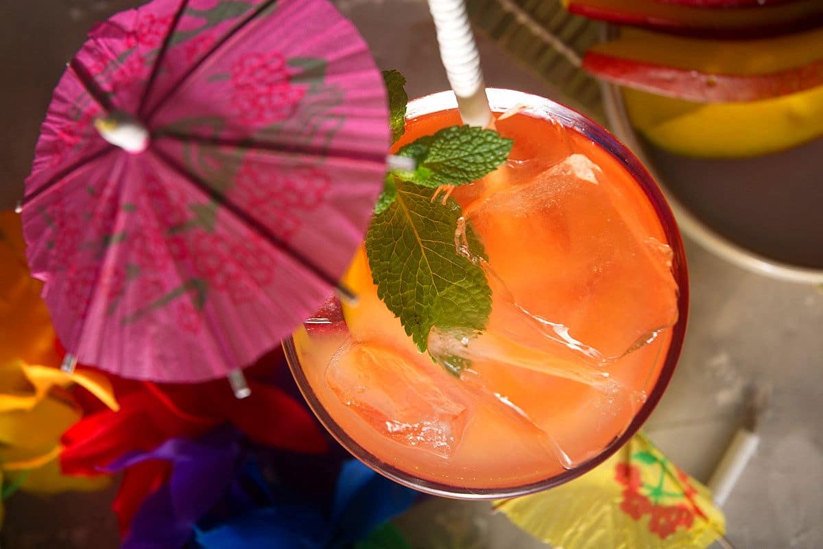 Top view of mai tai in a cocktail glass with pink umbrella, mango slice, mint sprig and straw.