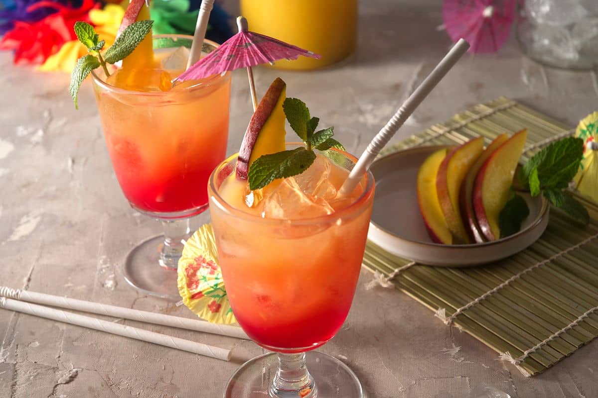 Mai tais in cocktail glasses with umbrella, mango slice, mint sprig and straw on concrete-style background.