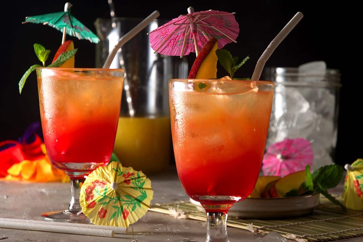 Mai tais in cocktail glasses with umbrella, mango slice, mint sprig and straw on black background.