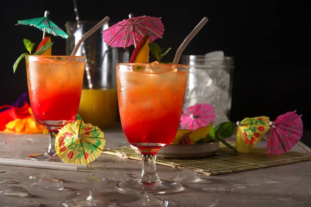 Mai tais in cocktail glasses with umbrella, mango slice, mint sprig and straw on black background.