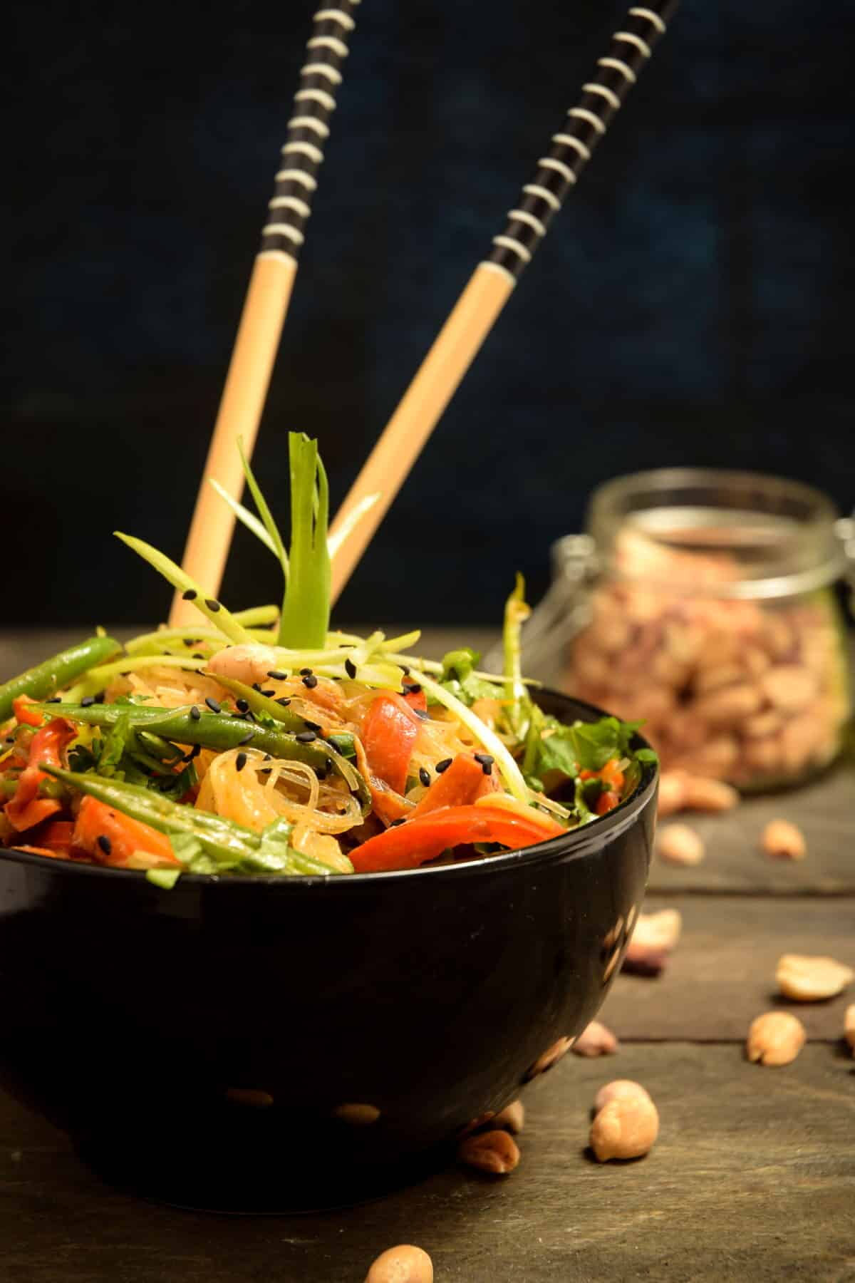 Vegan Pad Thai in a black bowl with chopsticks and peanuts in a jar in the background.