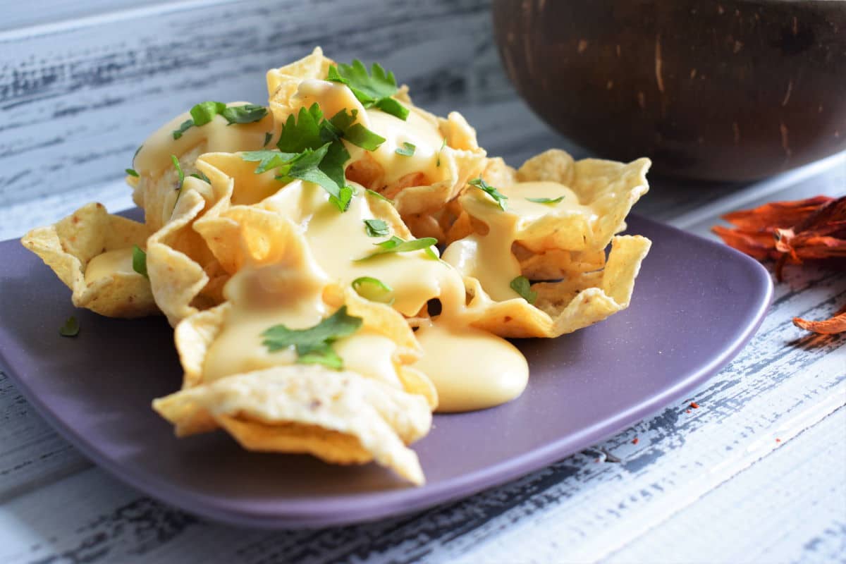 Tortilla chips with nacho cheese sauce on a purple plate, white wooden background.