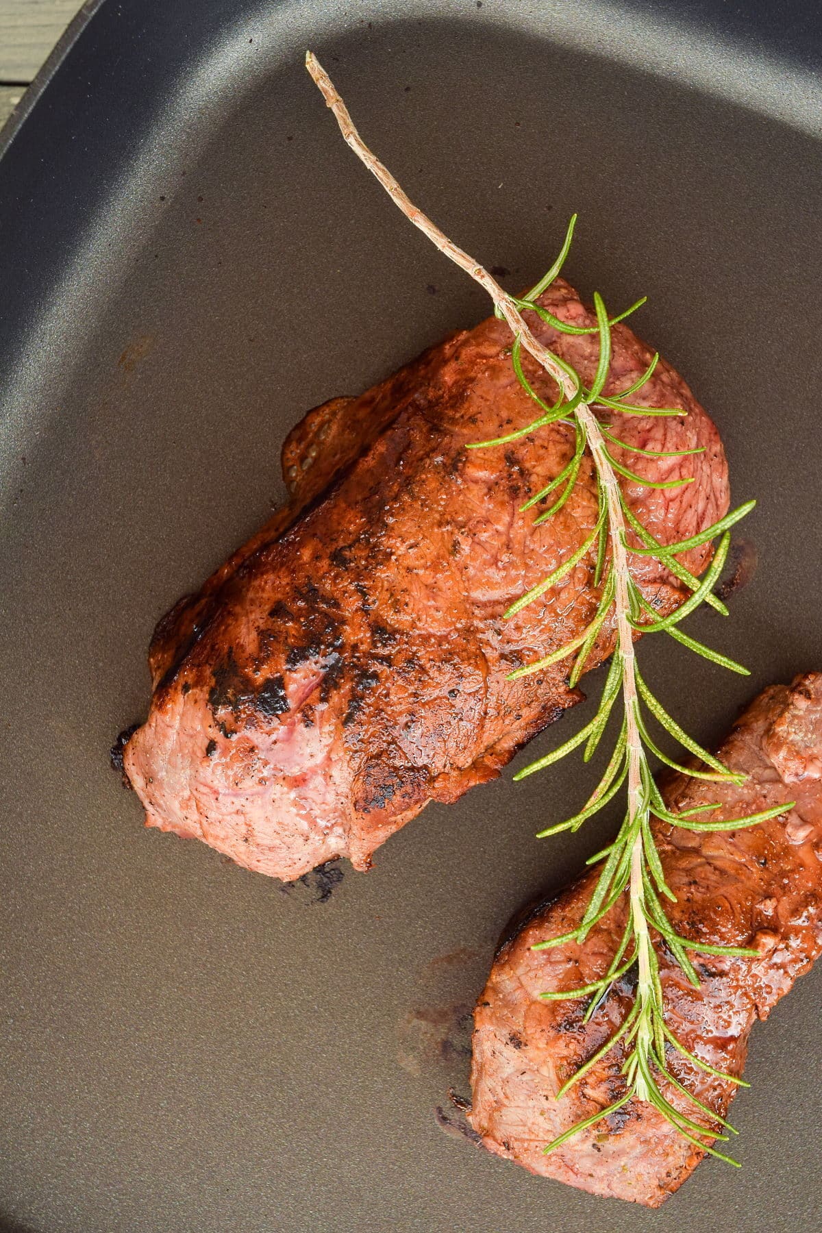 2 steaks in an electric skillet with a rosemary sprig on top.