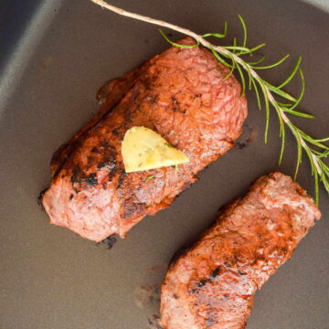 2 steaks in an electric skillet with butter and a rosemary sprig on top.