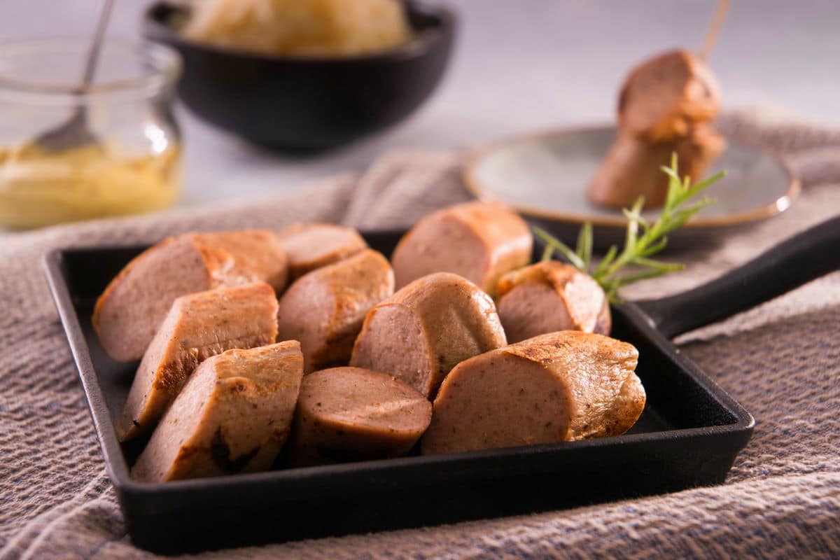 A cast iron pan filled with sliced sausages on a grey dish towel, sauerkraut and mustard in the background.