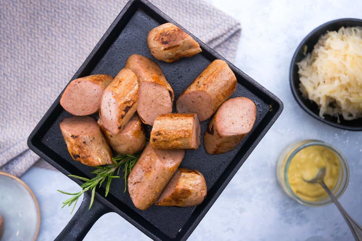 A cast iron pan filled with sliced sausage on a grey dish towel, sauerkraut and mustard on the side.