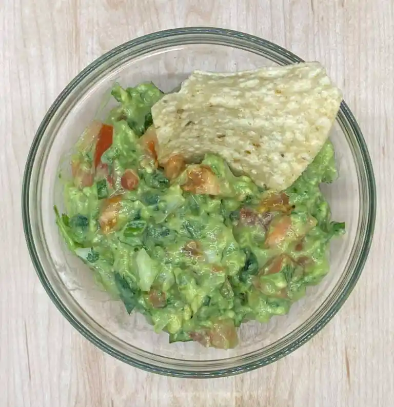 Guacamole with a tortilla chip in a glass bowl on wooden background.