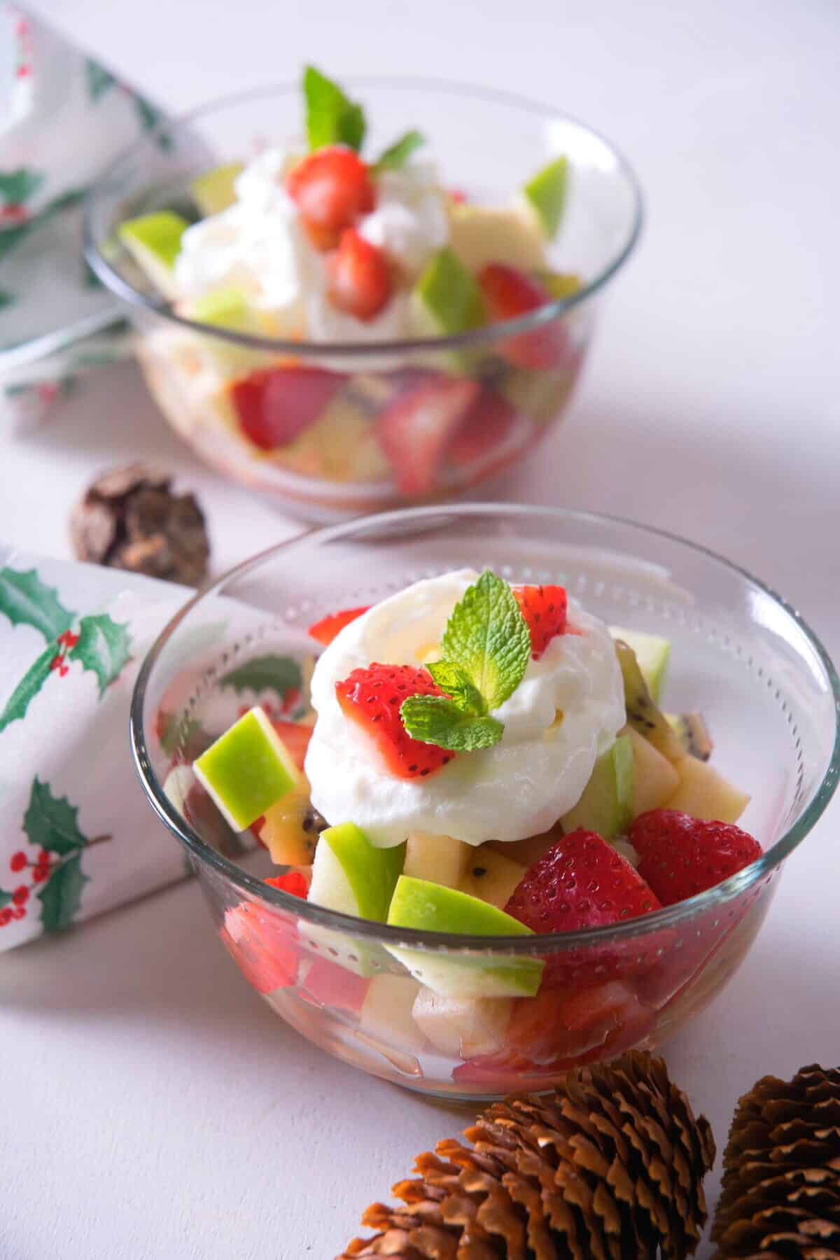 2 glass bowls with Christmas fruit salad. Pine cones and Christmas napkins on the side.
