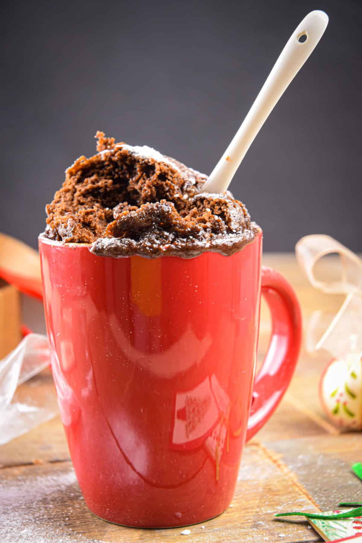 Chocolate mug cake in a red mug with a white spoon and Christmas decorations in the background.