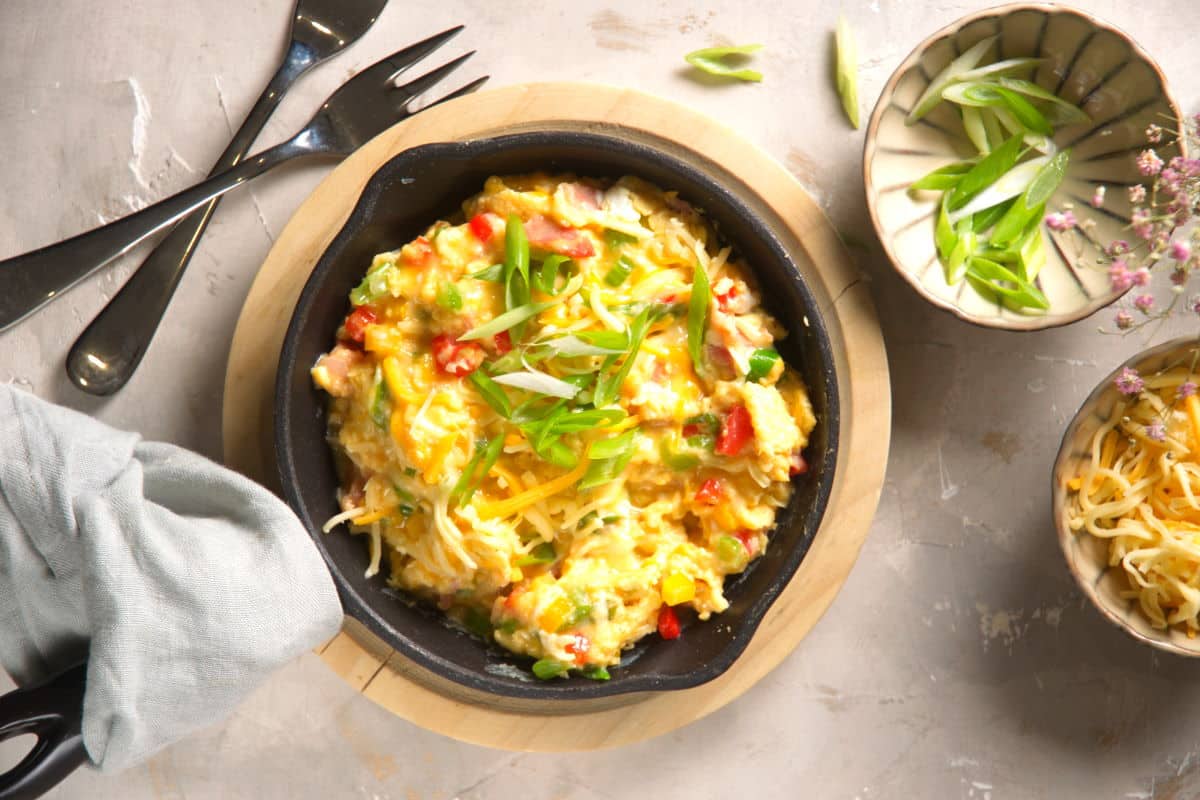 Scrambled eggs in cast iron pan with sliced green onions and grated cheese in small bowls on the side.