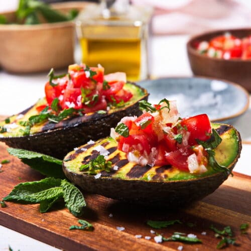 Close up of grilled, stuffed avocados on wooden cutting board.