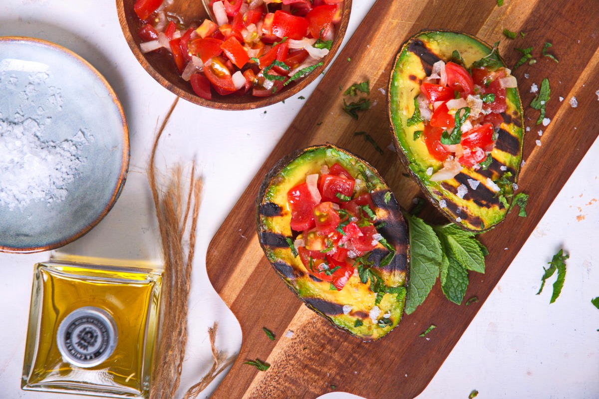 Top view of stuffed, grilled avocadoes on wooden cutting board with olive oil and salt on the side.