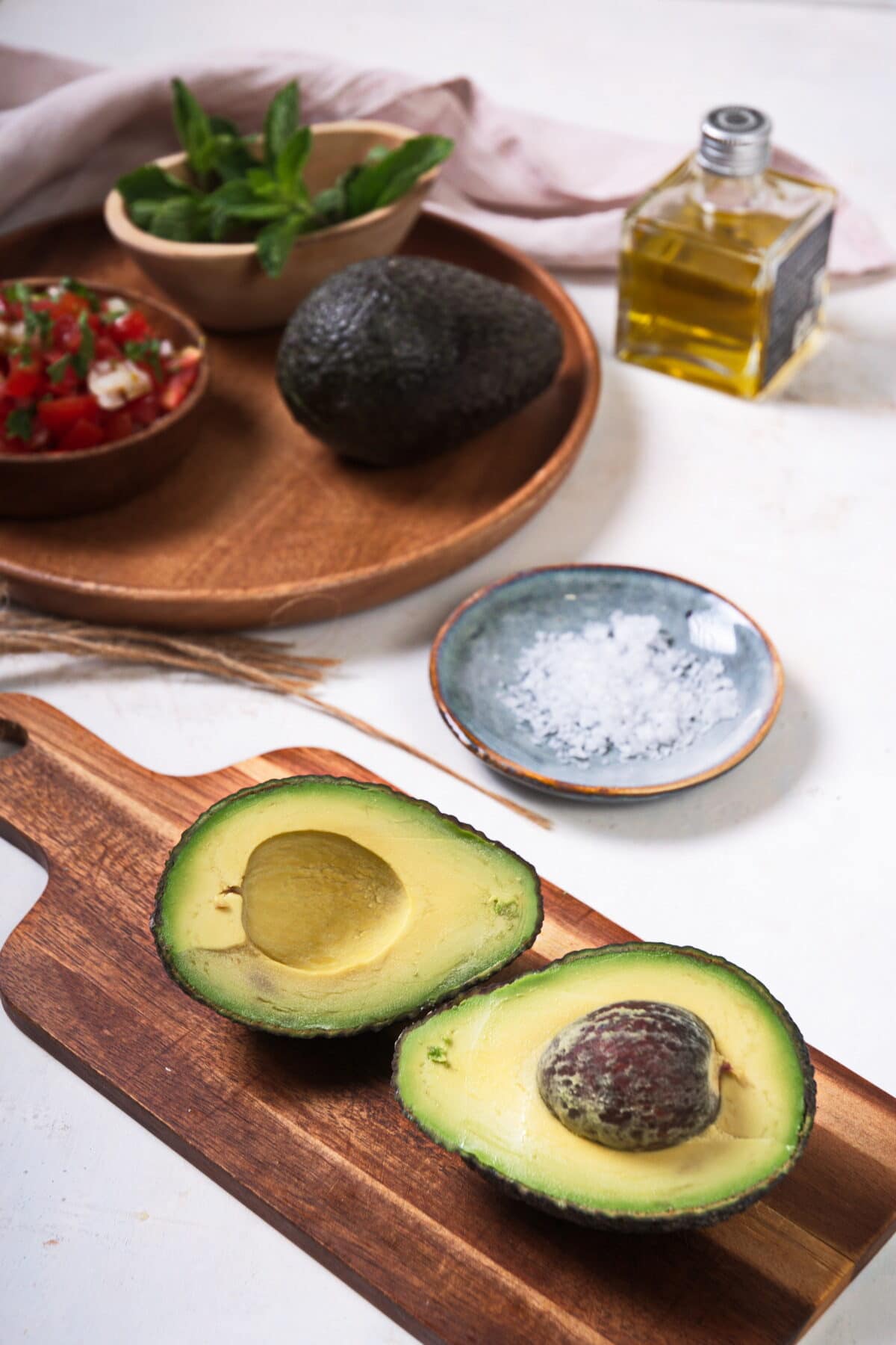 Sliced open avocado on wooden cutting board. Salt, olive oil and pico de gallo in the background.