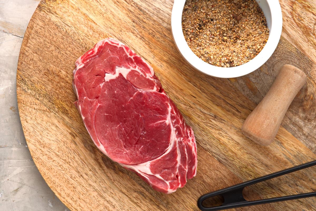 Raw bison ribeye steak on a wooden serving board with a bowl of spices.