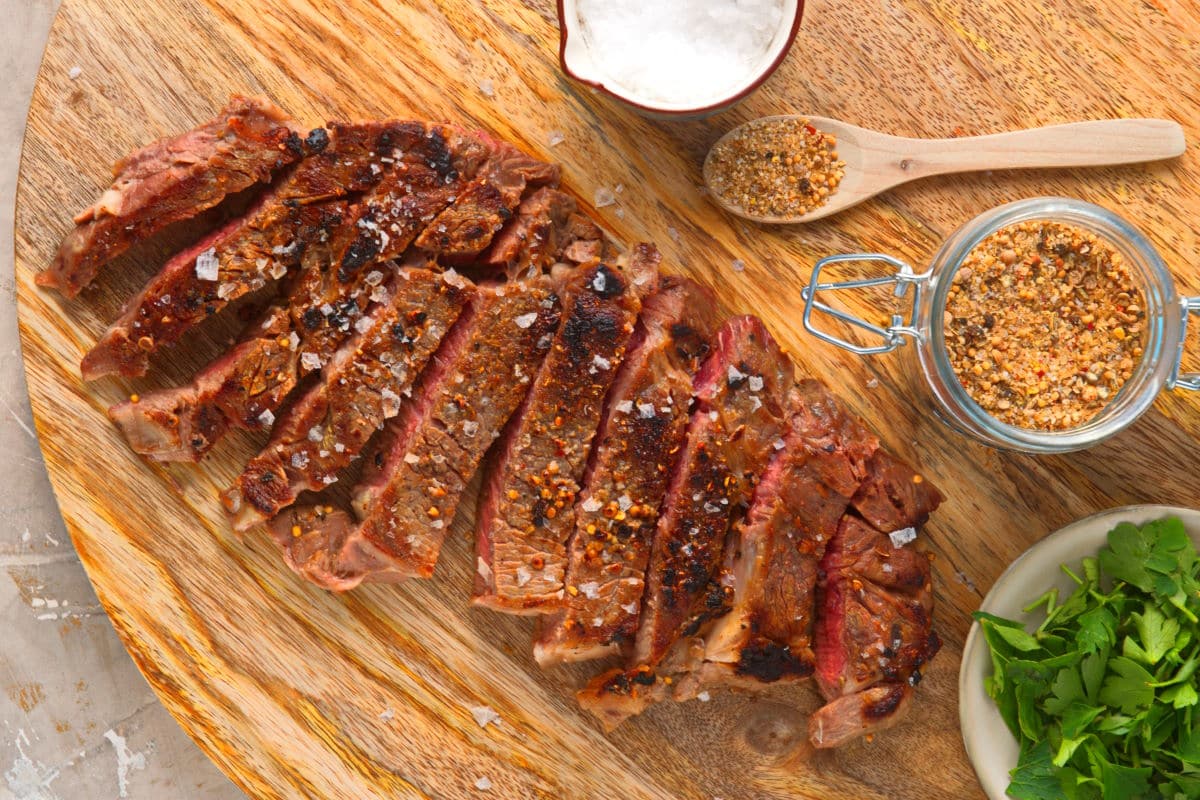 Grilled bison ribeye steak sliced on wooden serving board with spices on the side.