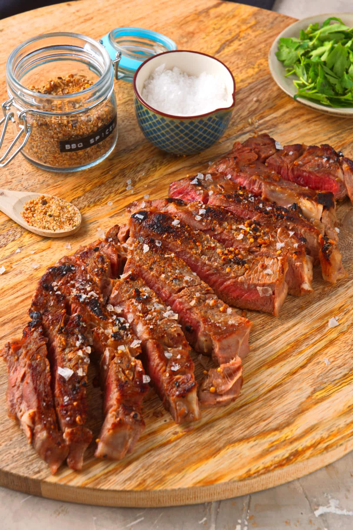 Grilled bison ribeye steak sliced on wooden serving board with spices on the side.