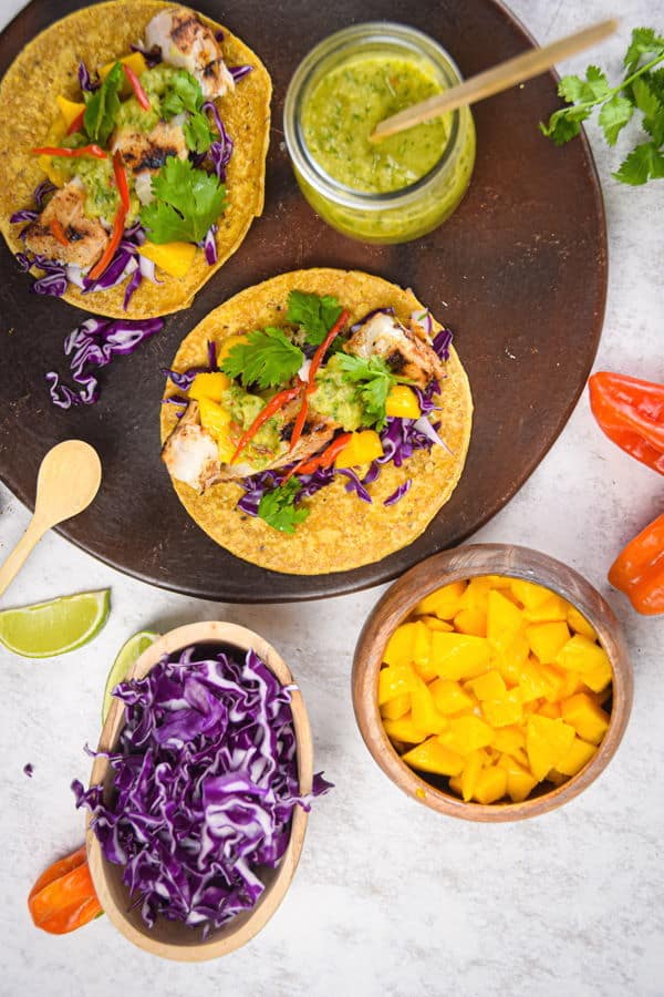 Grilled halibut tacos on brown wooden plate, salsa, cabbage, mangos and limes in the background.