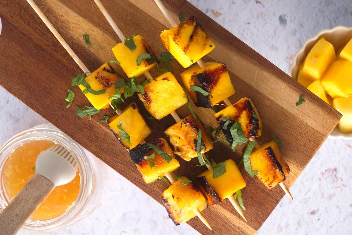 Grilled mango skewers on wooden serving board with fresh cilantro, marinade on the side.