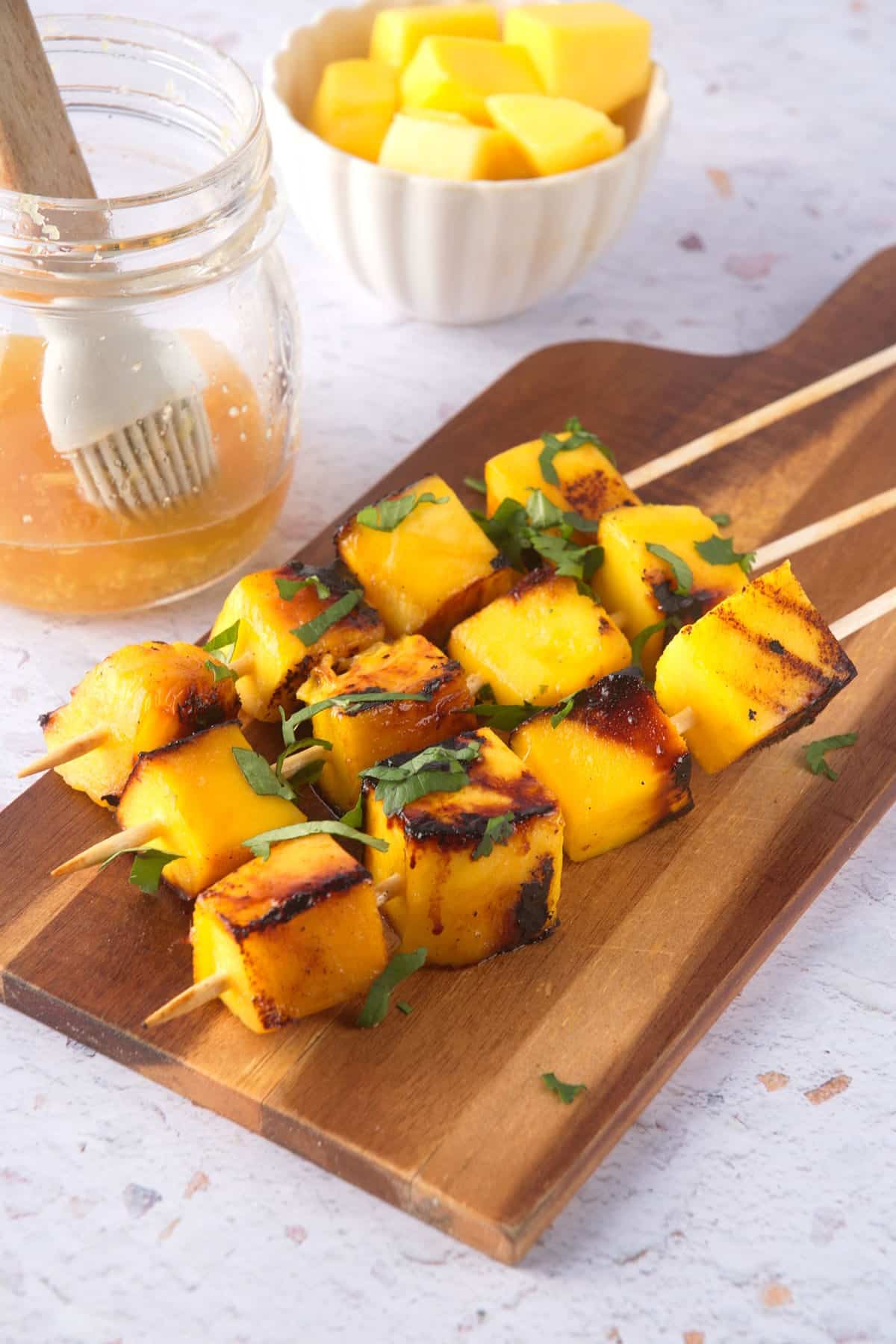 Grilled mango skewers on wooden serving board with fresh cilantro, marinade on the side.